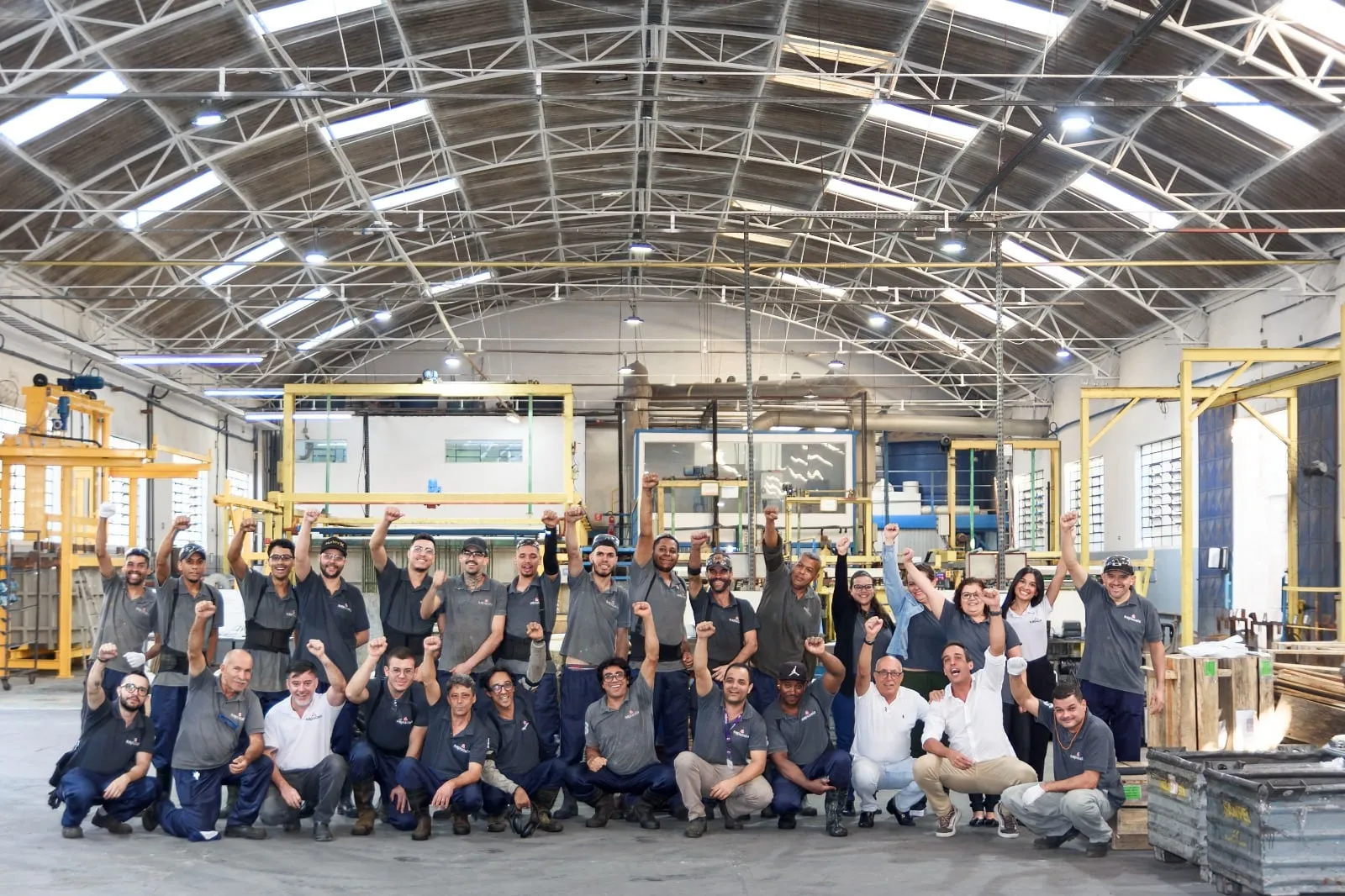 Equipe da Sapucaia reunida no galpão de produção, cerca de trinta pessoas com os braços erguidos sob a estrutura metálica do telhado