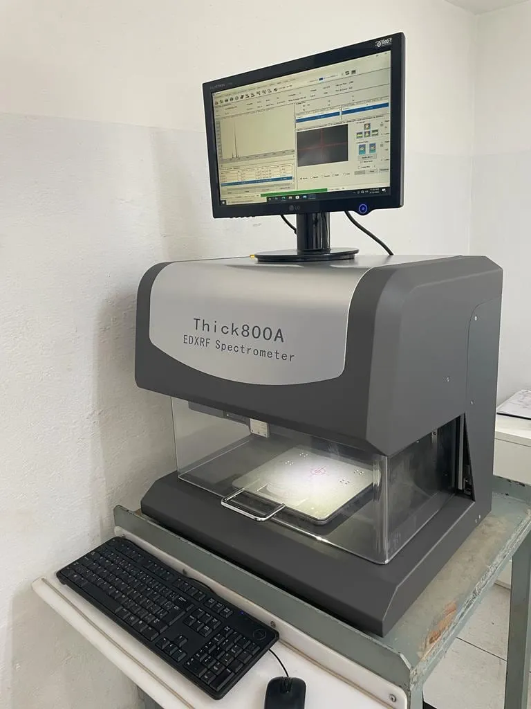 Espectrômetro de fluorescência de raios X Thick800A na bancada do laboratório, com o monitor exibindo o software de medição e a peça posicionada na câmara