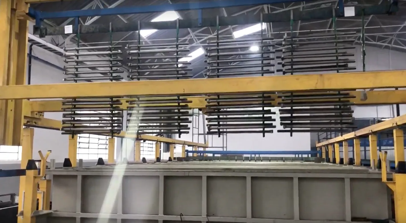 Interior da fábrica: pórtico amarelo e barramentos suspensos sobre a fileira de tanques da linha de banhos