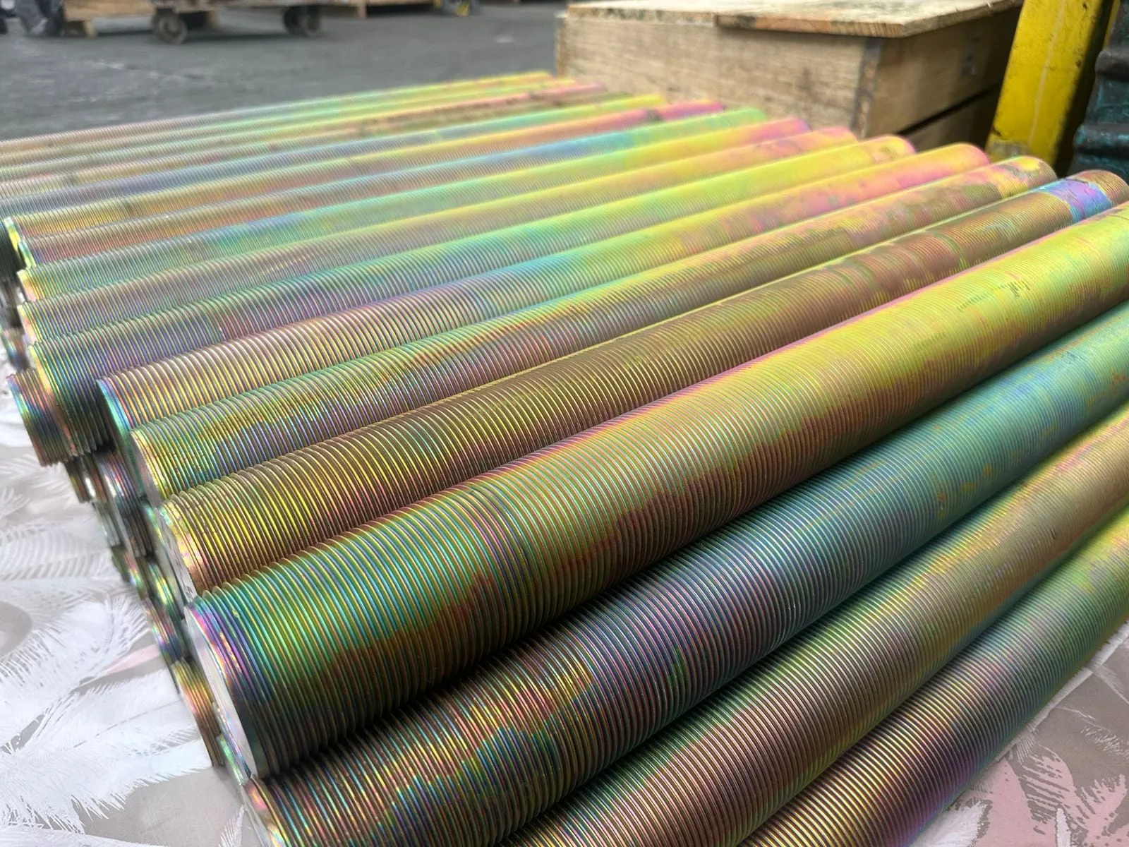 Barras roscadas empilhadas em pallet de madeira, com acabamento iridescente e o pórtico amarelo da linha ao fundo