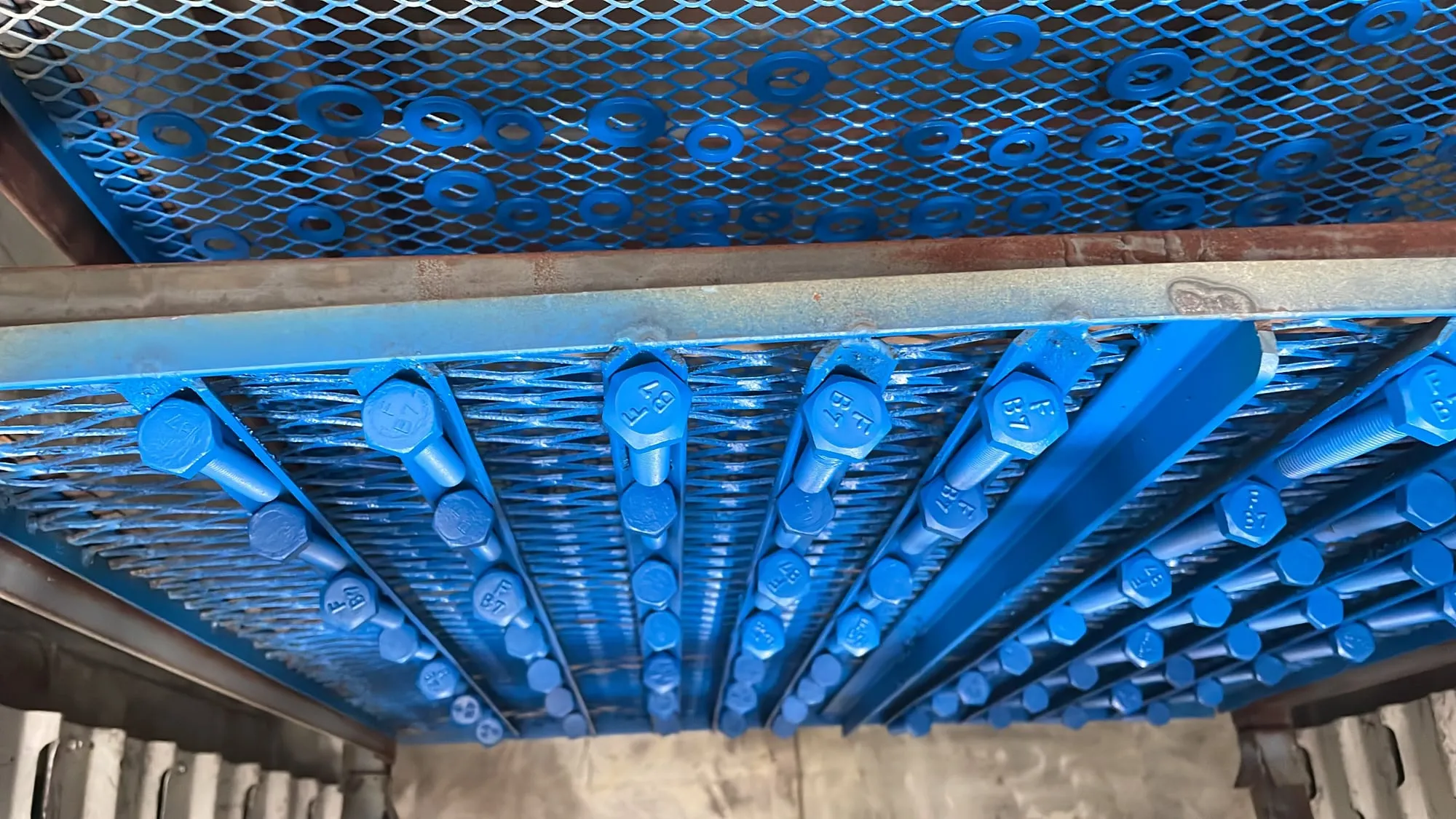 Parafusos azuis em prateleira de metal com fundo industrial. Superfície gradeada, arruelas soltas abaixo, clima de fábrica.