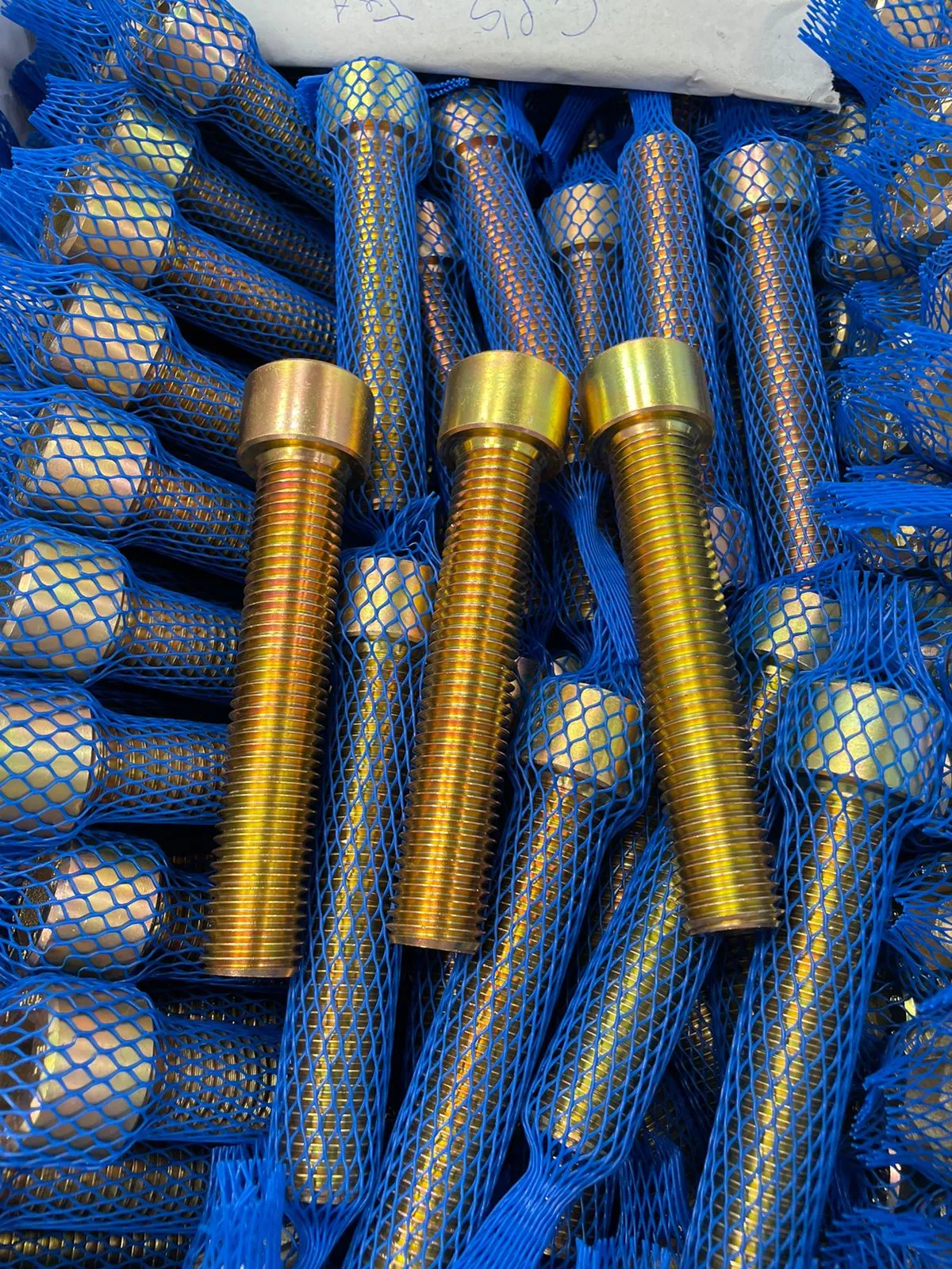 Parafusos allen de cabeça cilíndrica em acabamento amarelo-dourado iridescente, três em primeiro plano e o restante do lote ainda em malha azul de proteção